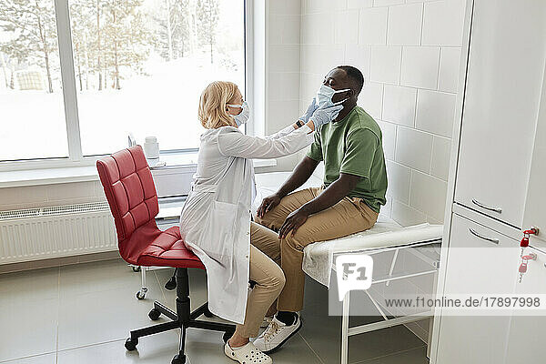 Doctor examining patient wearing protective face mask sitting on bed in clinic