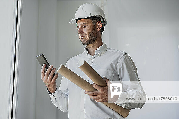 Mature engineer holding rolled up paper looking at smart phone in front of white wall