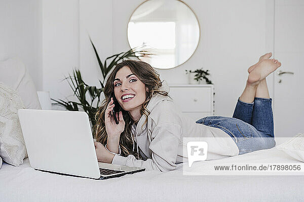 Smiling woman with laptop talking through phone lying on bed