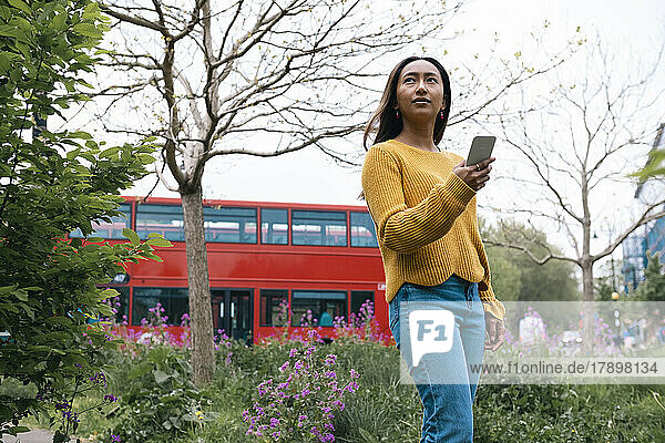 Nachdenkliche junge Frau mit Mobiltelefon steht im Park