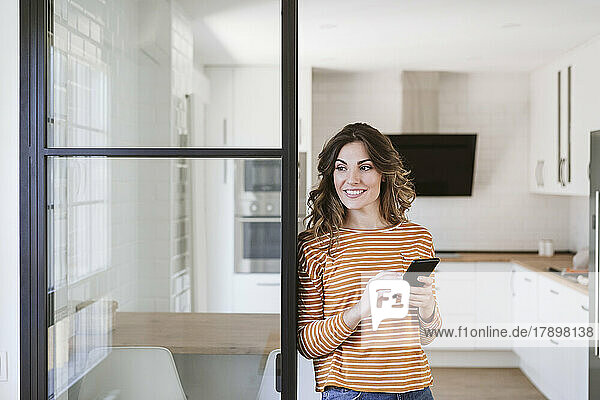 Smiling young woman holding mobile phone at a glass wall in kitchen