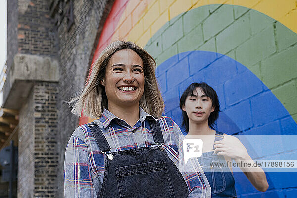 Happy young woman walking with friend by rainbow painted on wall