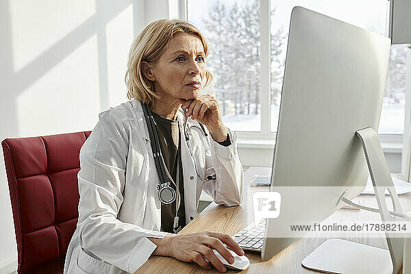 Doctor with hand on chin using desktop PC at desk in clinic