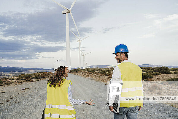 Smiling colleague gesturing at engineer holding solar panel at wind farm