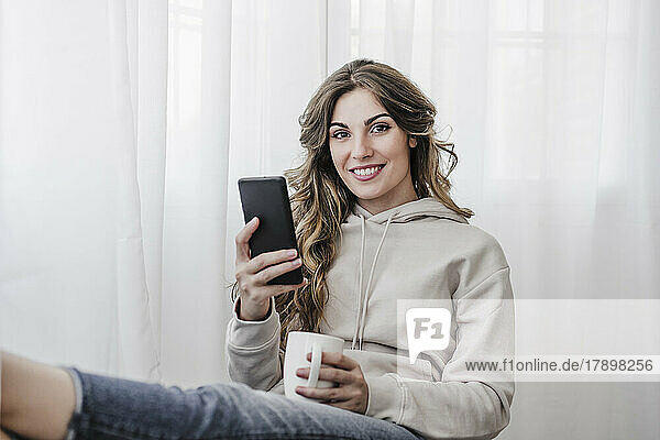 Lächelnde schöne Frau mit Kaffeetasse und Mobiltelefon sitzt zu Hause