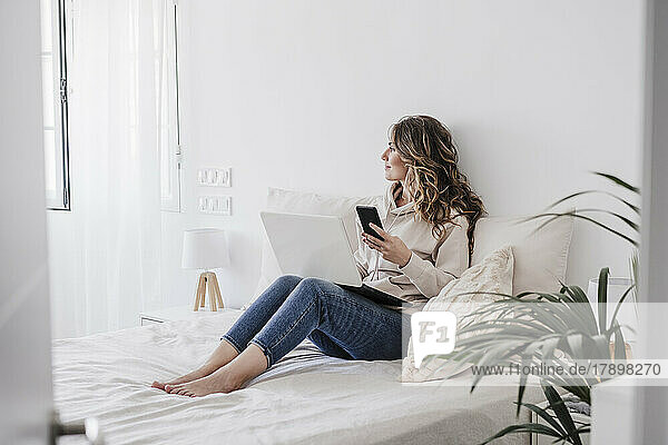 Nachdenkliche Frau mit Laptop und Telefon sitzt zu Hause im Bett