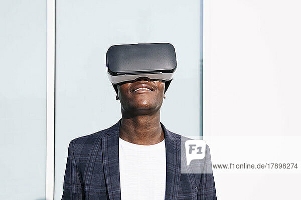 Smiling young man wearing VR glasses in front of wall