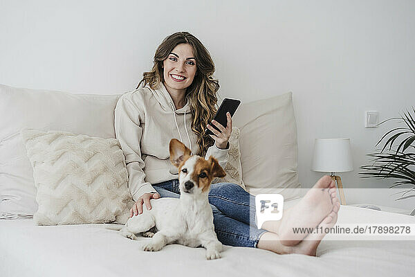 Smiling woman holding phone sitting by dog on bed at home