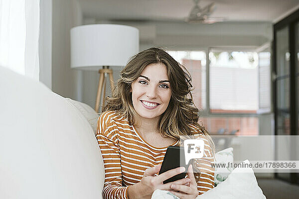 Portrait of smiling young woman with mobile phone on sofa at home
