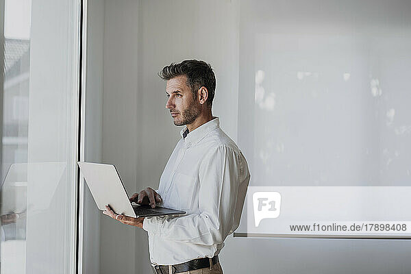 Thoughtful businessman holding laptop looking through window in office
