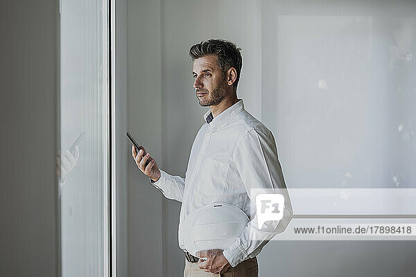 Thoughtful engineer holding hardhat and mobile phone looking through window in office