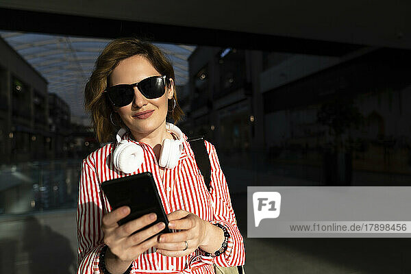 Lächelnde Frau mit Sonnenbrille und Smartphone im Einkaufszentrum