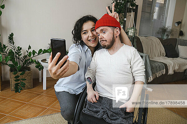 Happy woman with man in wheelchair taking selfie through mobile phone at home