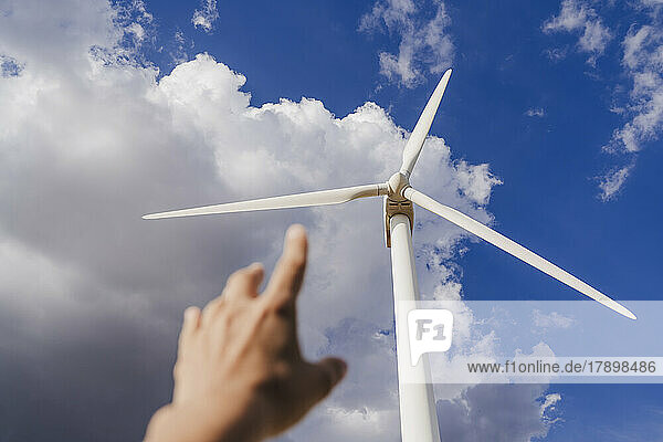 Hand of woman gesturing at rotor of wind turbine below clouds in sky