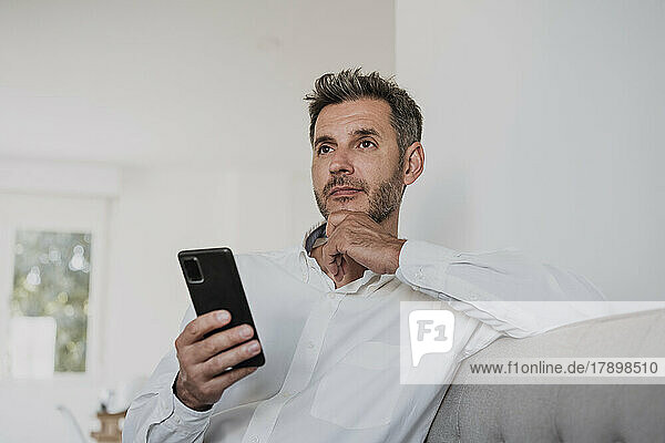 Thoughtful businessman holding smart phone on sofa in office