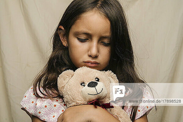 Trauriges Mädchen mit Teddybär vor beigem Vorhang