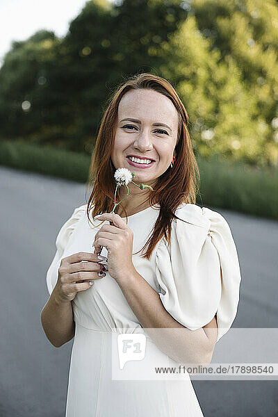 Smiling woman in white dress holding white flower at road