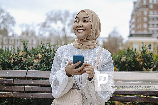 Happy young woman holding smart phone sitting on bench in park