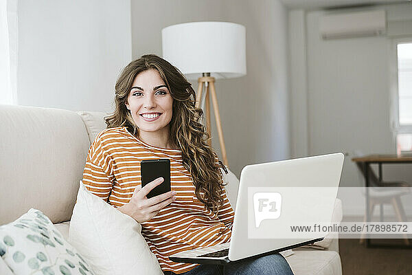 Smiling young woman with laptop and mobile phone on sofa at home