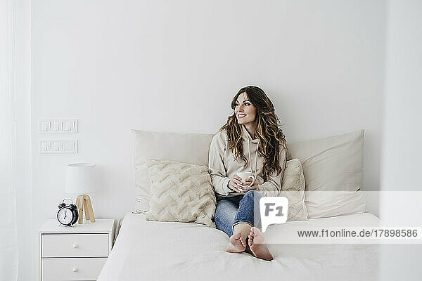 Lächelnde Frau mit Kaffeetasse sitzt zu Hause im Bett