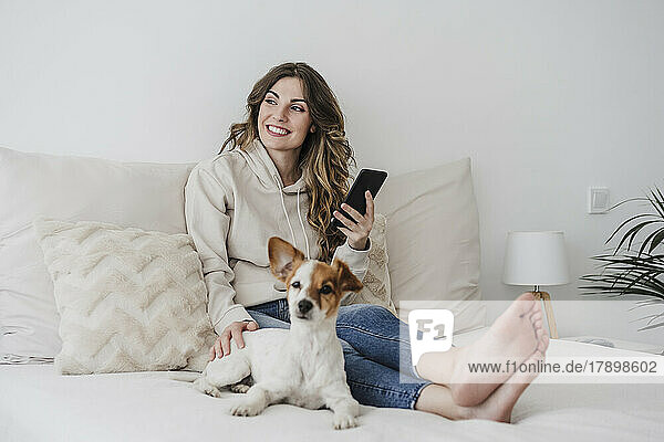 Lächelnde Frau mit Handy sitzt neben Hund im Schlafzimmer