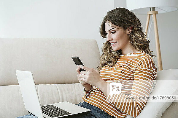 Smiling young woman with laptop using mobile phone on sofa at home