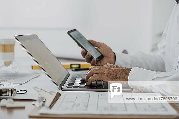 Hände des Architekten halten Mobiltelefon und Laptop am Schreibtisch