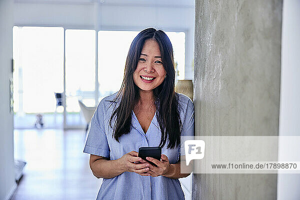 Happy woman with smart phone standing by wall at home
