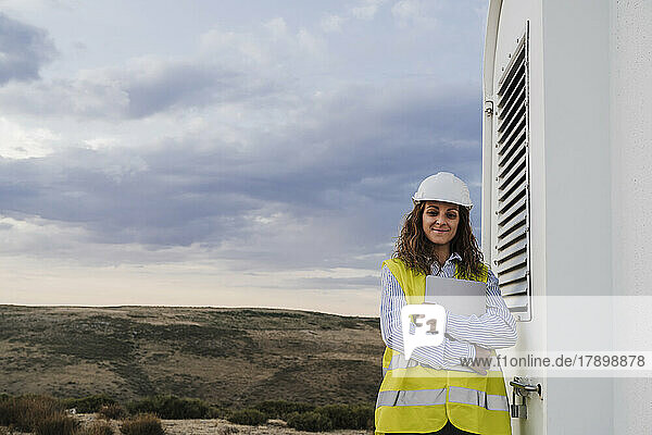 Confident engineer with laptop standing near door of wind turbine