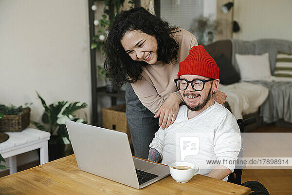 Smiling woman standing by boyfriend in wheelchair looking at laptop