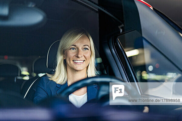 Happy businesswoman with blond hair driving car seen through windshield