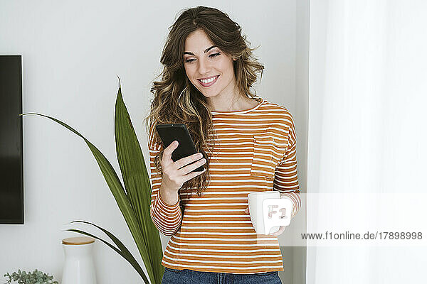 Smiling young woman holding coffee cup and using mobile phone