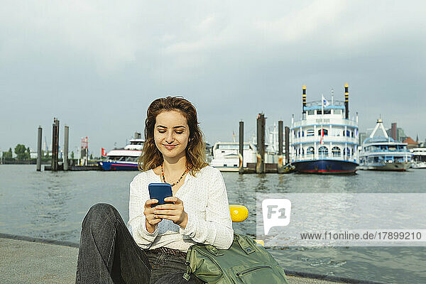 Smiling woman using smart phone sitting at Port of Hamburg  Germany