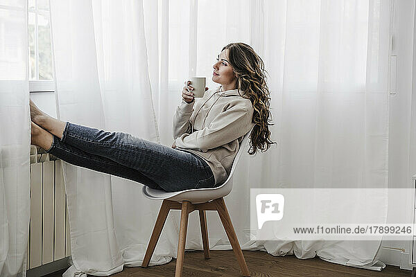 Junge Frau mit Kaffeetasse schaut durch das Fenster und sitzt auf einem Stuhl
