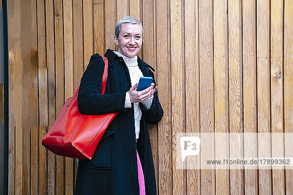 Happy woman with mobile phone leaning on wooden wall