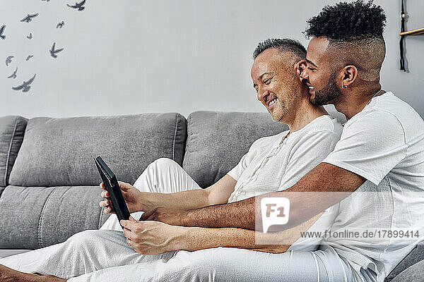 Smiling gay couple sharing tablet PC sitting on sofa at home