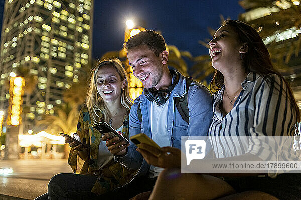 Cheerful women sitting by friend using smart phone at night
