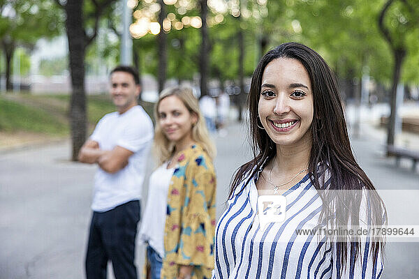 Lächelnde junge Frau mit langen Haaren steht vor Freunden im Park