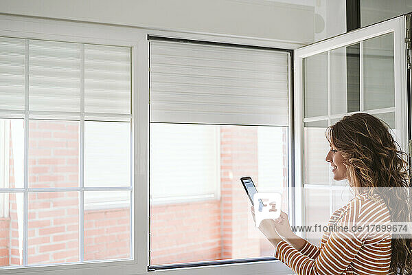 Young woman at home using mobile phone app to control blinds