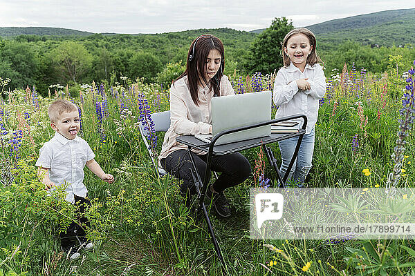 Playful children by mother working on laptop in meadow