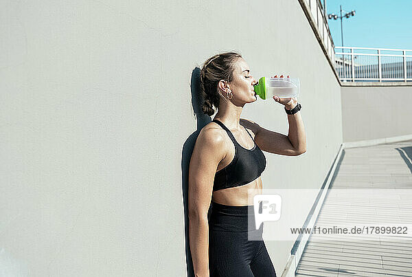 Woman drinking water from bottle leaning on wall