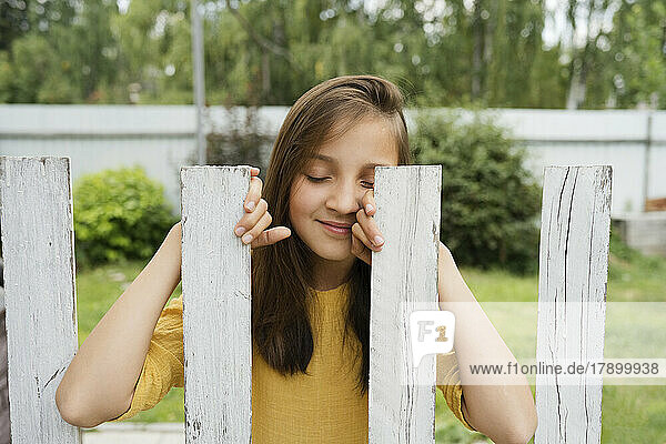 Smiling girl with eyes closed standing behind fence at back yard