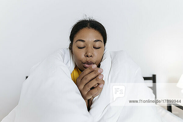 Freezing young woman sitting on the bed wrapped in a blanket rubbing hands