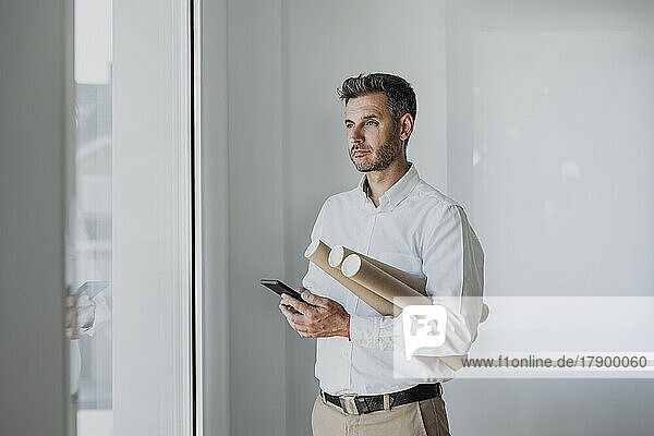 Thoughtful architect holding mobile phone and rolled up paper looking through window in office
