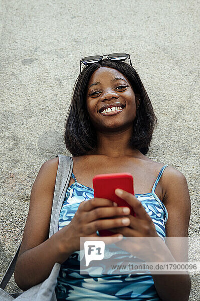 Smiling woman wearing sunglasses holding mobile phone in front of wall