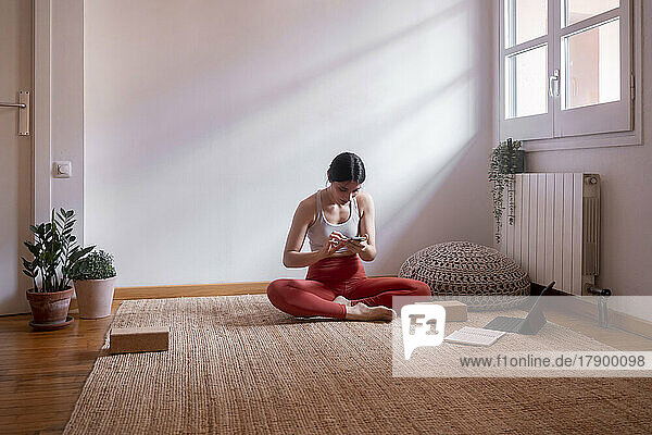 Young woman using smart phone by laptop sitting on carpet floor at home