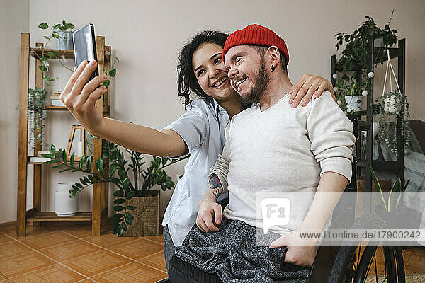 Smiling woman with boyfriend in wheelchair taking selfie through mobile phone at home