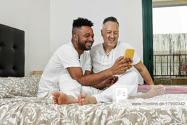 Gay couple sharing mobile phone sitting on bed at home