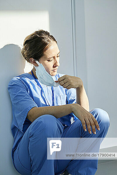 Krankenschwester entfernt Schutzmaske  während sie im Krankenhaus an der Wand hockt