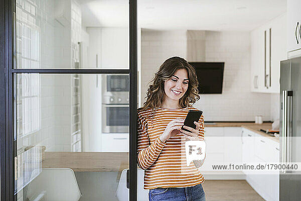 Smiling young woman using mobile phone at a glass wall in kitchen